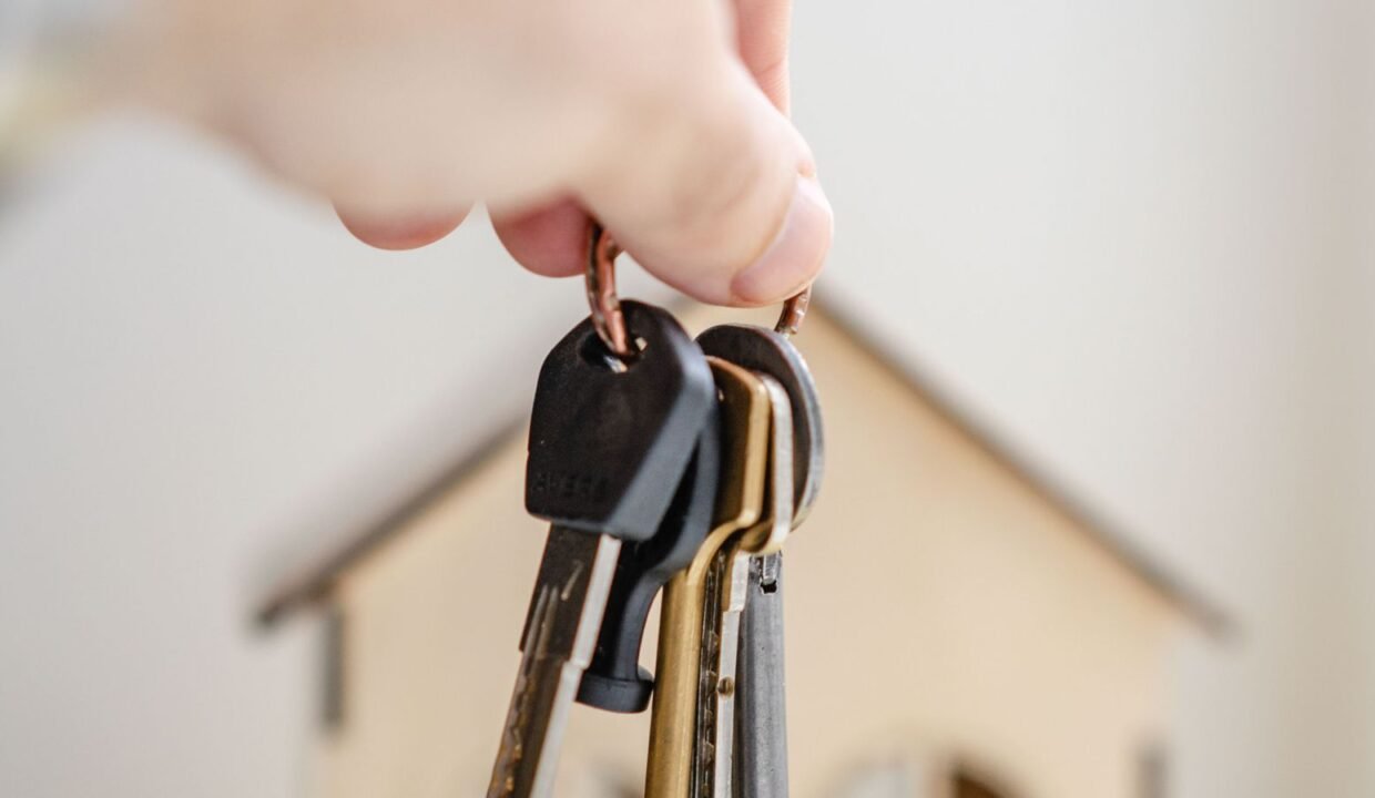 Hand holding house keys beside a miniature wooden house